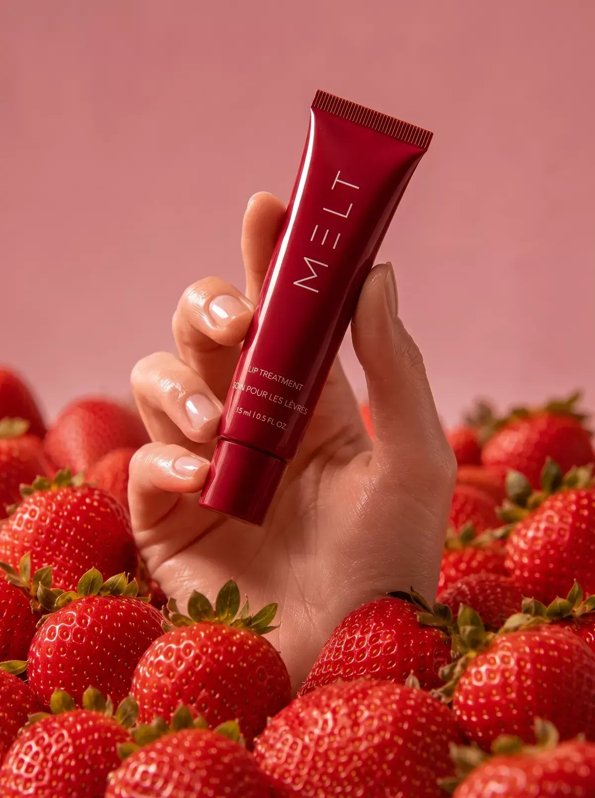 A hand holds the red MELT lip treatment tube above a bed of strawberries.